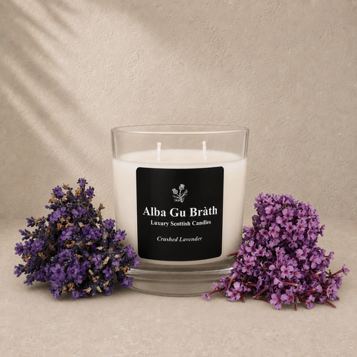 A white scented candle in a clear glass jar with a black label reading 'Alba Gu Bréth Luxury Scottish Candles' and 'Crushed Lavender.' The candle is styled on a beige surface with dried purple lavender flowers placed on both sides against a textured beige backdrop.