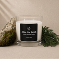 A clear glass jar candle with white wax and a black label reading 'Alba Gu Bréth Luxury Scottish Candles Forest Mist,' styled on a beige surface with a mossy rock and green pine sprigs against a textured beige backdrop.