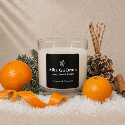 A glass jar candle with a black label reading 'Alba Gu Bréth Luxury Scottish Candles' is styled with oranges, orange peels, a pine cone, cinnamon sticks, and evergreen branches on white faux snow against a beige textured backdrop.