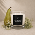 A white scented candle in a clear glass jar with a black label reading 'Alba Gu Bréth Luxury Scottish Candles', styled on a beige surface with a green pear and small white flowers against a textured beige wall.