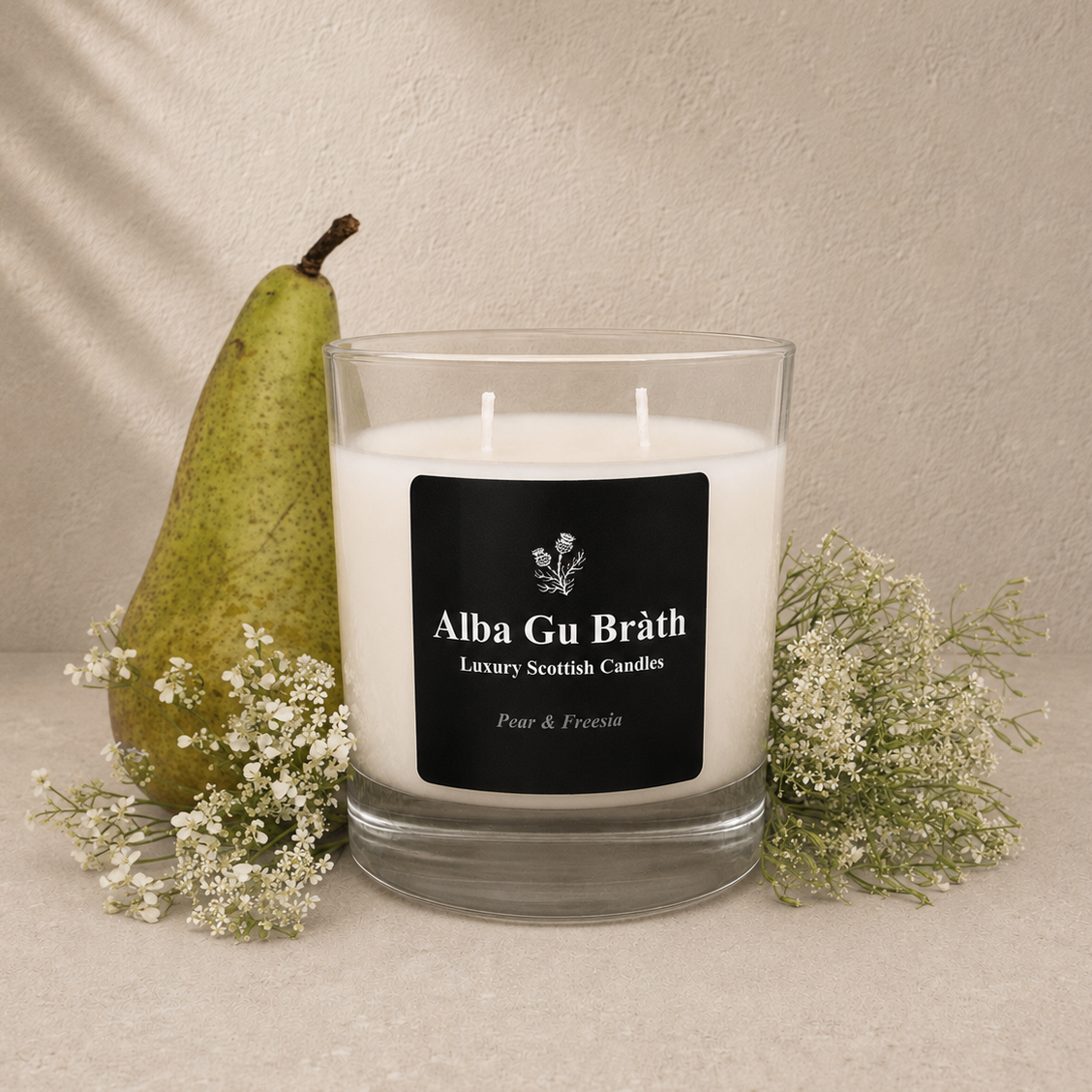 A white scented candle in a clear glass jar with a black label reading 'Alba Gu Bréth Luxury Scottish Candles', styled on a beige surface with a green pear and small white flowers against a textured beige wall.